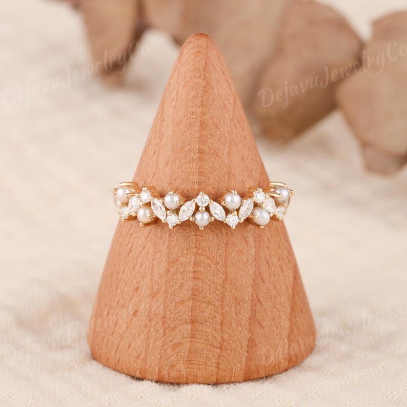 May include: A gold ring with a floral pattern, set with pearls and clear gemstones. The ring is displayed on a wooden cone. The design features alternating pearls and marquise-cut gemstones, showcasing the ring's intricate details and craftsmanship.
