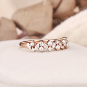 May include: Rose gold ring with a delicate design of pearls and marquise-cut and round gemstones. The ring has a floral pattern, with the gemstones and pearls arranged in a repeating motif. The ring is set on a white surface.