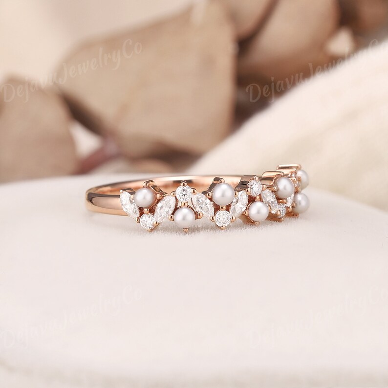 May include: Rose gold ring with a delicate design of pearls and marquise-cut and round gemstones. The ring has a floral pattern, with the gemstones and pearls arranged in a repeating motif. The ring is set on a white surface.