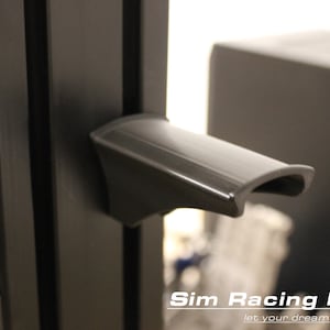 Könnte beinhalten: Nahaufnahme eines schwarzen Kunststoffgriffs, der an einem schwarzen Metallrahmen befestigt ist. Der Griff hat eine geschwungene Form und eine glatte Oberfläche. Der Text "Sim Racing Parts let your dreams come true" ist am unteren Rand des Bildes sichtbar.