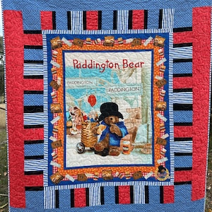 Könnte beinhalten: Eine rote, schwarze und blaue Patchwork-Decke mit einem zentralen Panel, das Paddington Bear zeigt. Das Panel zeigt Paddington Bear, der auf einer Bank mit einem Koffer und einem roten Ballon sitzt. Der Text "Paddington Bear" ist in Weiß auf dem Panel gedruckt.