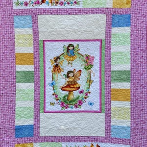 以下が含まれることがあります： ピンクと白のキルトで、中央のパネルにはキノコの上に妖精が描かれた幻想的なイラストが施されています。パネルは緑、黄色、青の縁取りで囲まれています。キルトは花柄と小さな鳥で飾られています。