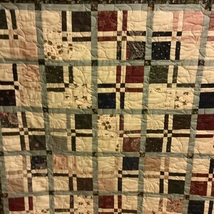 Könnte beinhalten: Eine Steppdecke mit Patchwork-Muster, bestehend aus Quadraten aus verschiedenen Stoffen in Braun-, Beige-, Rot- und Blautönen. Die Steppdecke ist mit einem Muster aus geschwungenen Linien gesteppt.
