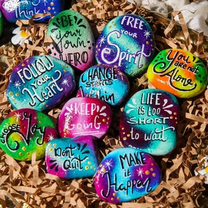 May include: A selection of hand-painted stones with motivational messages in different colours. The stones display phrases such as "Follow Your Heart," "Keep Going," and "Make it Happen." White daisy accents are scattered around the stones.