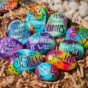 May include: A collection of painted rocks with inspirational messages, including "Escape the Ordinary", "Expect Nothing Appreciate Everything", "Life is too Short", "Push Your Limits", "It Will Be", "You are Strong", "Stay Fab", "Be Kind", and "Stay Curious".