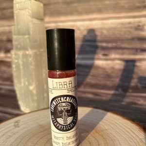 May include: A small, clear glass roll-on bottle with a black cap, labeled "Libra Spell Oil" by Lovewitch Creations. The label features a two-faced figure and the words "Beauty, Balance, Self, Relationships." A white, textured crystal and a wooden slice are in the background.