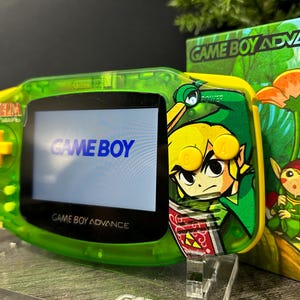 May include: A green Game Boy Advance handheld console with a Zelda-themed design. The screen displays "GAME BOY" and "GAME BOY ADVANCE". The console is next to a box with a colorful illustration of characters.