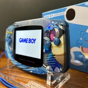 May include: A Game Boy Advance handheld console with a custom design featuring a blue and white character illustration. The screen displays "GAME BOY". The console is on a clear acrylic stand, with a blue lanyard attached.