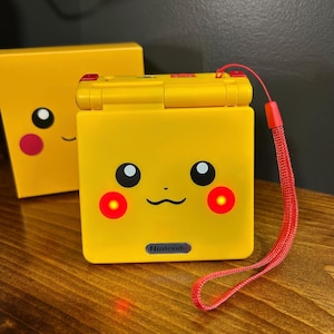 May include: Yellow Nintendo Game Boy Advance SP handheld game console with a Pikachu design. Features a red wrist strap, black eyes, red cheeks, and a black curved mouth. The Nintendo logo is visible.