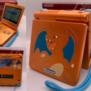May include: Orange Game Boy Advance SP handheld game console with a Charizard graphic. The console has a blue strap and buttons. The box is visible in the background. The screen displays the word "GAMEBOY".