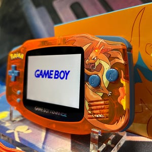May include: Orange Game Boy Advance handheld console with a Charizard design. The screen displays "GAME BOY" in blue. The console has blue buttons and an orange wrist strap. A matching box is visible in the background.
