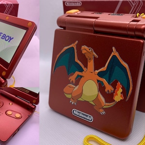 May include: A metallic red Game Boy Advance SP console with a yellow strap. The console features a screen displaying "GAME BOY" and a sticker of the Pokémon character Charizard. The Nintendo logo is visible.