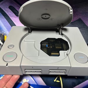 May include: A light gray PlayStation 1 console with the disc tray open. The console has a reset button, an open button, and two controller ports. Cables and a hand are visible.