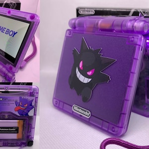 Puede incluir: Consola Game Boy Advance SP púrpura transparente con una pegatina de Gengar. La pantalla muestra "GAME BOY". La consola tiene una correa de muñeca púrpura y se ve un cartucho de juego. La caja del fondo dice "Nueva pantalla de luz frontal".