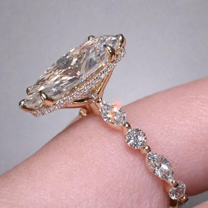 Könnte beinhalten: Ein goldener Verlobungsring mit einem großen Diamanten im Marquiseschliff. Der Ring hat kleinere Diamanten entlang des Bandes und um den Hauptstein herum. Das Design ist elegant und funkelnd.