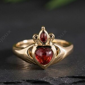 Peut inclure: Bague dorée ornée d'une pierre rouge en forme de cœur. La bague présente une couronne au-dessus du cœur et des mains qui l'étreignent. L'ensemble est posé sur un fond sombre. Bijou de style Claddagh.