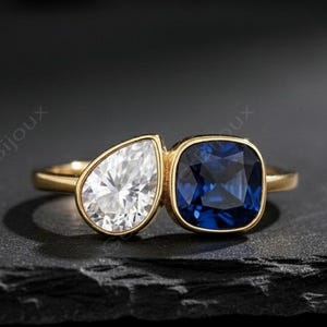 以下が含まれることがあります： 2つの宝石をあしらったゴールドリング。洋梨型の白い石と、正方形にカットされた深い青色の石が特徴です。暗い背景にセットされ、宝石の輝きを際立たせています。