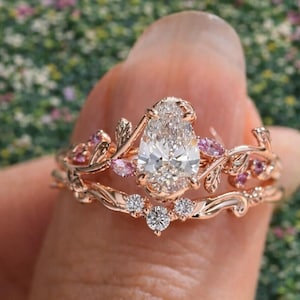 May include: Rose gold engagement ring set featuring a pear-cut diamond and floral detailing. The band includes pink gemstones and a matching band with small diamonds, creating a nature-inspired design.