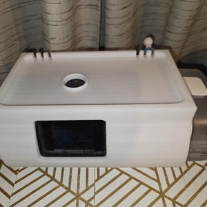 May include: A white rectangular device with a dark opening and a circular hole on top. The device has a lid and is next to a metallic component with a white button. The device is on a gold and white patterned surface.