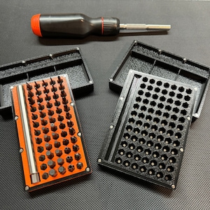 Puede incluir: Un organizador de puntas negro y naranja con una variedad de puntas de destornillador. El organizador rectangular tiene un cierre magnético. Un destornillador negro y rojo está encima del organizador.