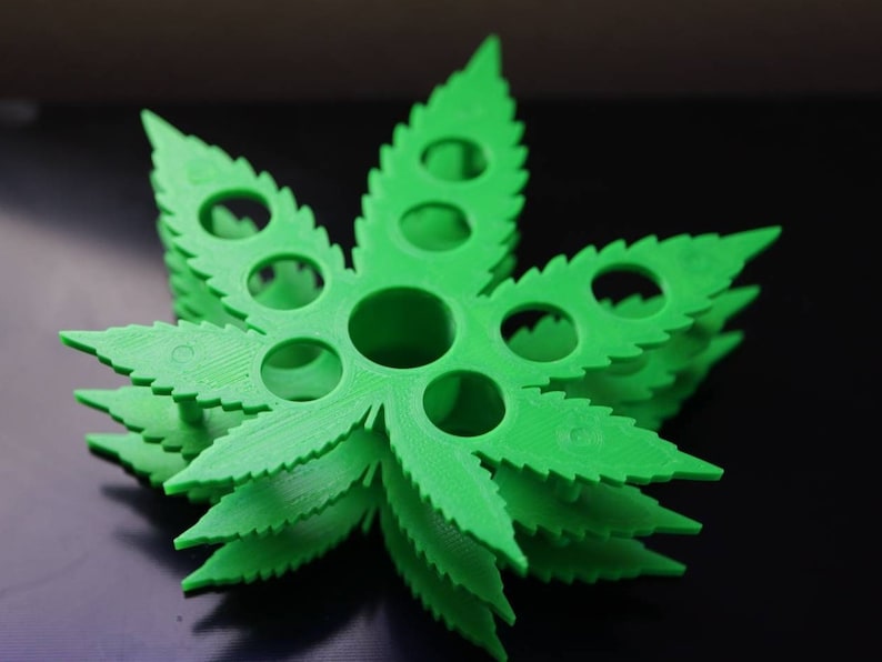 K&ouml;nnte beinhalten: Ein gr&uuml;ner, kunststoffgefertigter Marihuana-Blatt-f&ouml;rmiger Halter mit mehreren runden L&ouml;chern. Der Halter ist &uuml;bereinander gestapelt und erzeugt so einen dreidimensionalen Effekt.