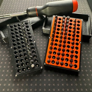May include: Two black and orange cases filled with a variety of screwdriver bits. The cases are made of plastic and have a honeycomb pattern. A black and red screwdriver is lying on top of the cases.