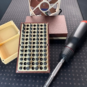 May include: A close-up of a set of screwdriver bits organised in a grid pattern. The bits are arranged in a brown and beige plastic case. A silver screwdriver with a black handle is lying on a black and white patterned surface.