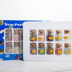 Puede incluir: Un conjunto de pequeños frascos de vidrio llenos de piedras semipreciosas, incluyendo labradorita, cuarzo rosa y granate. Cada frasco tiene un tapón de corcho y una etiqueta. La caja está etiquetada como "SEMI-PREC".
