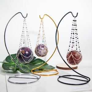 Puede incluir: Tres esferas de cristal colgando de soportes metálicos. Cada esfera está encerrada en una jaula de cuentas y tiene un esquema de color diferente. Las esferas se muestran sobre un fondo blanco.