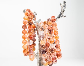 May include: A set of three bracelets made with red, orange, and brown gemstone beads. The bracelets are displayed on a silver tree-shaped stand.