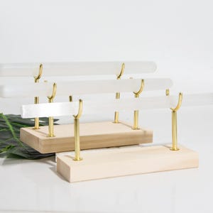 Pode incluir: Um conjunto de três suportes de exibição de cristal de selenite. Cada suporte apresenta uma base de madeira retangular, suportes de metal dourado e uma barra horizontal de cristal de selenite. Os suportes estão numa superfície branca.