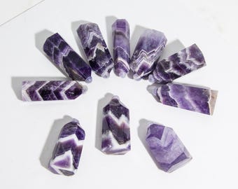 May include: Ten amethyst crystal wands with a chevron pattern in shades of purple and white. The wands are arranged in a circle on a white surface.
