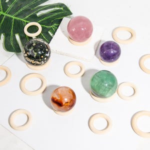 May include: A collection of six polished crystal spheres in various colours, including green, pink, purple, and brown, displayed on wooden rings. The spheres are arranged on a white surface.