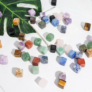 Pode incluir: Uma coleção de pequenos cubos de pedras preciosas, de cores variadas e polidas. Os cubos são feitos de diferentes tipos de pedras, incluindo ametista, quartzo rosa, olho de tigre e aventurina verde. Os cubos estão espalhados sobre uma superfície branca.