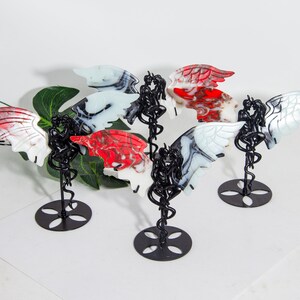 May include: Four black metal stands with a swirling design, each holding a resin sculpture of two figures embracing. The figures have wings and are painted in a mix of white, red, and black.