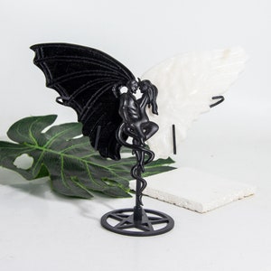 May include: Black and white sculpture of a couple embracing, with wings, on a pentagram stand. The sculpture is made of resin and is a decorative piece.