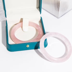 Puede incluir: Dos pulseras de cuarzo rosa de color rosa. Las pulseras están en una caja verde azulada con un cierre dorado.