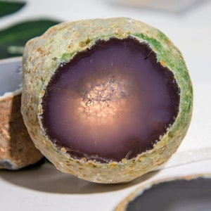 May include: A close-up of a geode with a rough, textured exterior in shades of green and tan. The interior reveals a smooth, polished surface in a deep purple hue, with a sparkling, crystalline center. The geode is a natural stone formation.