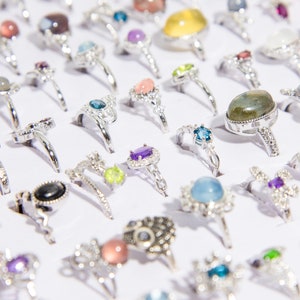 May include: A display of silver rings with various colored gemstones. The rings are arranged in rows on a white surface. The gemstones include blue, green, purple, and black.