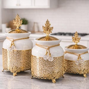 Op de afbeelding: Drie decoratieve witgouden potten met bladvormige finials. De potten hebben een witte keramische bovenkant en een gouden filigraan basis. Elke pot is versierd met een gouden ketting en kristallen accenten. De potten worden tentoongesteld op een marmeren aanrecht.