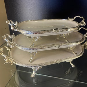 May include: Three tiered serving trays with silver-tone rims and ornate handles. The trays are oval shaped and have a white ceramic base.