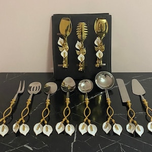 May include: Set of gold and silver serving utensils with white flower accents. The utensils include a serving spoon, a serving fork, a ladle, a butter knife, and a cake server.