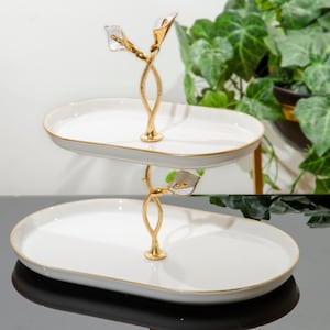 Puede incluir: Bandeja de servir de cerámica blanca de dos niveles con un diseño floral dorado. La bandeja es de forma ovalada y tiene un borde dorado.