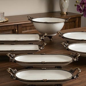 Può includere: Una collezione di piatti da portata bianchi con finiture e manici decorativi color bronzo. Il set comprende vassoi ovali, rettangolari e rotondi, oltre a una grande ciotola, tutti appoggiati su piccoli piedini. I piatti sono disposti su un tavolo di legno.