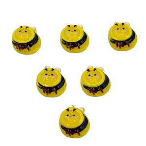 May include: Six small, yellow bee-shaped charms with black stripes and brown accents. Each charm has a smiling face and small, clear wings. The charms are arranged on a white background.