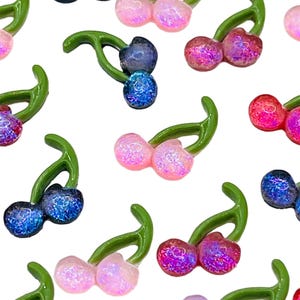 May include: A pattern of glittery cherry charms in various colors, including pink, red, blue, and purple. Each charm has a green stem and is scattered across a white background. These charms are ideal for crafting or jewelry making.