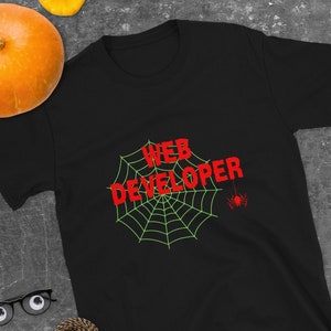 May include: Black t-shirt featuring the text "WEB DEVELOPER" in red, set against a green spider web design and a small red spider. The shirt is on a dark grey surface with Halloween decorations.
