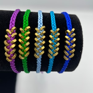 May include: Five braided bracelets with gold-coloured metal beads. The bracelets are in shades of purple, green, blue, turquoise, and royal blue.