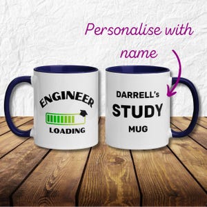 May include: White ceramic mug with a blue rim and handle. The mug has a black and green progress bar with the text "Engineer Loading" and a graduation cap. The other side of the mug has the text "Darrell's Study Mug".