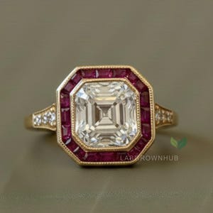 May include: A gold ring featuring a large, square, clear central gemstone, encircled by a halo of small, red stones. The ring has a vintage design with small accent stones on the band.